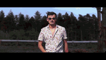 a man wearing sunglasses and a floral shirt stands in front of trees