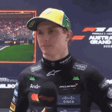 a man wearing a hat and a black shirt is standing in front of a screen that says formula 1 austria grand prix
