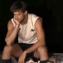 a man wearing a white tank top and black shorts is sitting down with his hand on his chin .