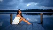 a woman in a white dress is sitting on a pier overlooking a lake .