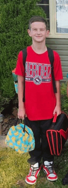 a young boy in a red shirt is standing in front of a house holding a backpack and lunch bag .
