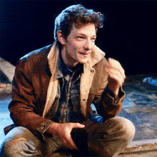 a man wearing a brown jacket and a denim shirt is sitting on the floor