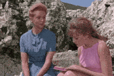 two women are sitting on a rock reading a book