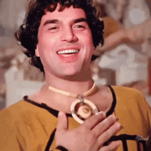 a man wearing a yellow shirt and a necklace is smiling and holding his hand to his chest