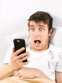 a man is laying in bed looking at a cell phone with a surprised look on his face .