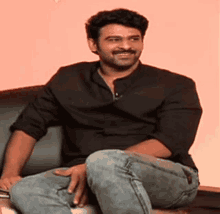 a man in a black shirt and jeans is sitting on a couch and smiling .