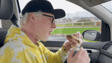 a man in a yellow hoodie and black hat is eating a hot dog