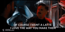 a man with a mustache is talking to another man in a dark room while holding a cup of coffee .