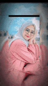 a woman wearing glasses and a hijab looks at the camera with her hands on her face