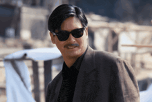 a man with a mustache wearing sunglasses and a suit looks at the camera