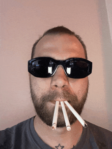 a man with a beard wearing sunglasses has three cigarettes sticking out of his mouth