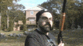 a man with a beard and glasses holds a shotgun in front of a house
