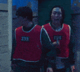 a man wearing a red vest with the number 334 on it stands next to another man wearing a green vest with the number 124 on it