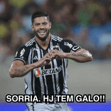 a soccer player wearing a black and white striped jersey is smiling and making a heart shape with his hands .