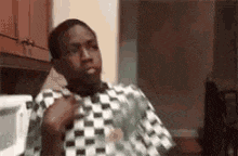 a man is sitting in a kitchen wearing a checkered shirt and making a funny face .