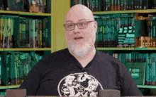 a bald man with glasses and a beard is standing in front of a bookshelf filled with books