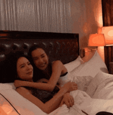 two women are hugging each other on a bed with a lamp in the background