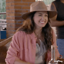 a woman wearing a hat and a pink jacket is smiling while sitting at a table with a bottle of wine .