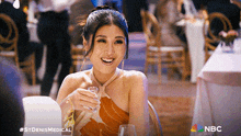 a woman is sitting at a table with a glass of wine in her hand and a nbc logo behind her