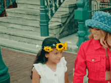 a little girl wearing a sunflower crown stands next to a woman wearing a louis vuitton hat