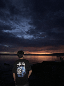 a man wearing a stussy shirt stands in front of a body of water