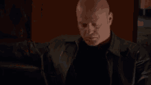 a bald man in a leather jacket and black shirt is sitting in a chair .