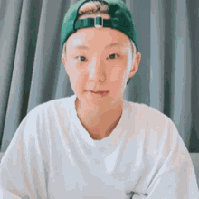a young man wearing a green baseball cap and a white t-shirt is sitting in front of a curtain .