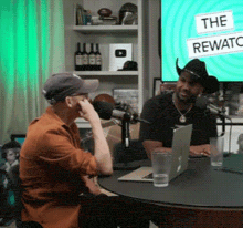 two men are sitting at a table with a sign that says the rewards