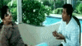 two men are sitting on a couch talking to each other in front of a swimming pool .