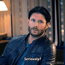 a man with a beard wearing a leather jacket is sitting in a chair and says seriously .