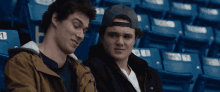 two young men are sitting in a stadium watching a game and one has the number 1 on his seat