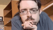 a man with glasses and a mustache is sitting in front of a shelf .
