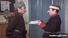 two men in military uniforms are standing next to each other and one is holding a pan .
