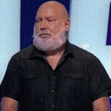 a bald man with a beard wearing a black shirt is standing in front of a sign .