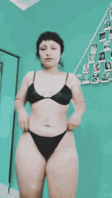 a woman in a black bikini stands in front of a green wall with pictures hanging on it