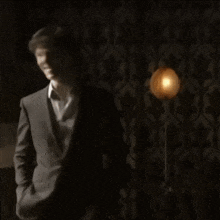 a man in a suit stands in front of a lamp and says brilliant yesss