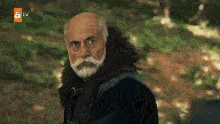 a man with a beard and a fur coat is standing in the woods looking at the camera .