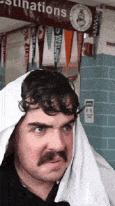 a man with a mustache and a towel around his head stands in front of a sign that says destinations