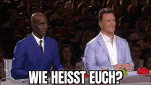 two men are sitting at a table in front of a crowd and one of them is asking the question .