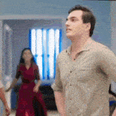 a man in a tan shirt stands in a room with a woman in a red dress in the background