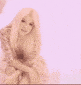 a woman with long blonde hair is sitting on a box with her legs crossed and a pink background .