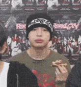 a man wearing a beanie is holding a slice of pizza in front of a poster that says roadway