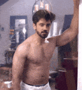 a shirtless man leans against a wall with his arm up