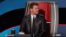 a man in a leather jacket is sitting in a chair with a nbc logo in the background