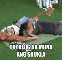 two women are laying on the grass and one is holding a book