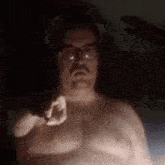 a shirtless man with glasses is pointing at the camera in the dark .