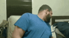 a man with a beard is sitting on a bed wearing a blue shirt and a blue shirt .
