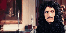 a man in a wig and mustache is sitting in a chair in front of a painting .