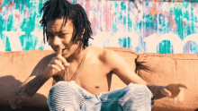 a shirtless man with dreadlocks sits on a couch in front of a graffiti wall