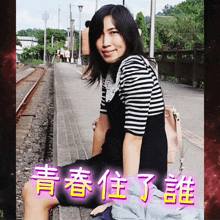 a woman in a striped shirt sits on a bench in front of a train track